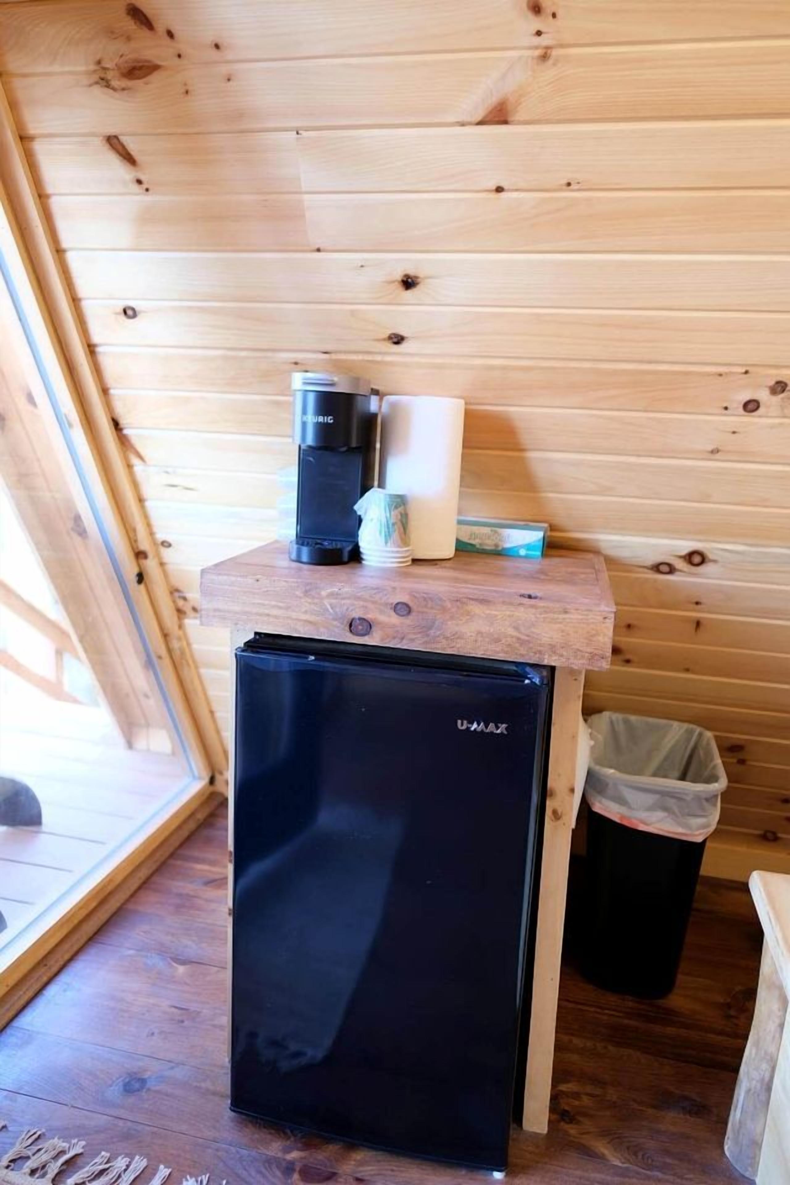 Ett kompakt köksområde med ett svart U-MAX-kylskåp, en kaffebryggare och en papperskorg.