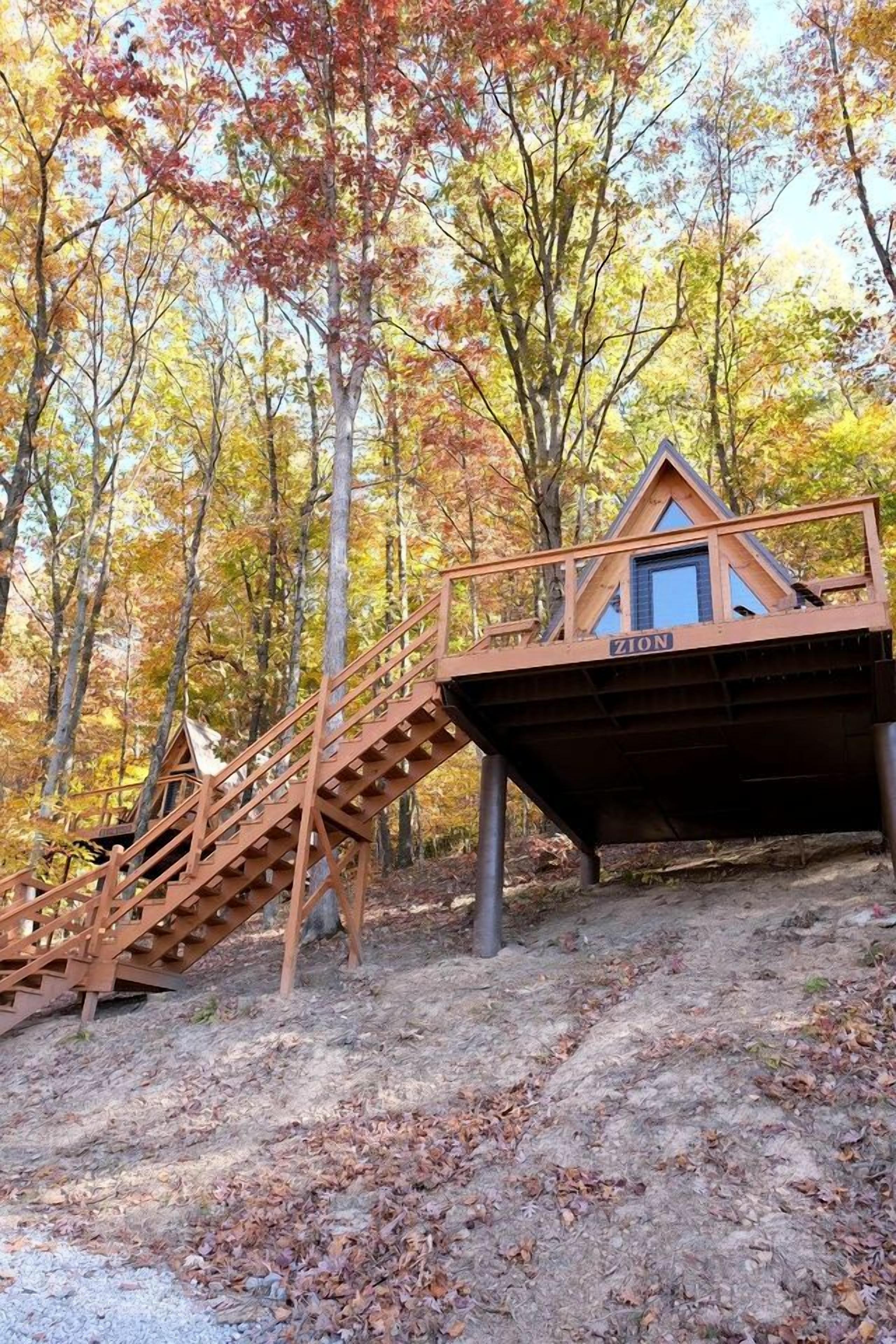 A wooden cabin with a staircase leading up to it, surrounded by trees with autumn foliage.