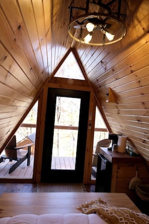 Una cabaña de madera con techo a dos aguas, una cama con una manta estampada, una mesita y una silla.