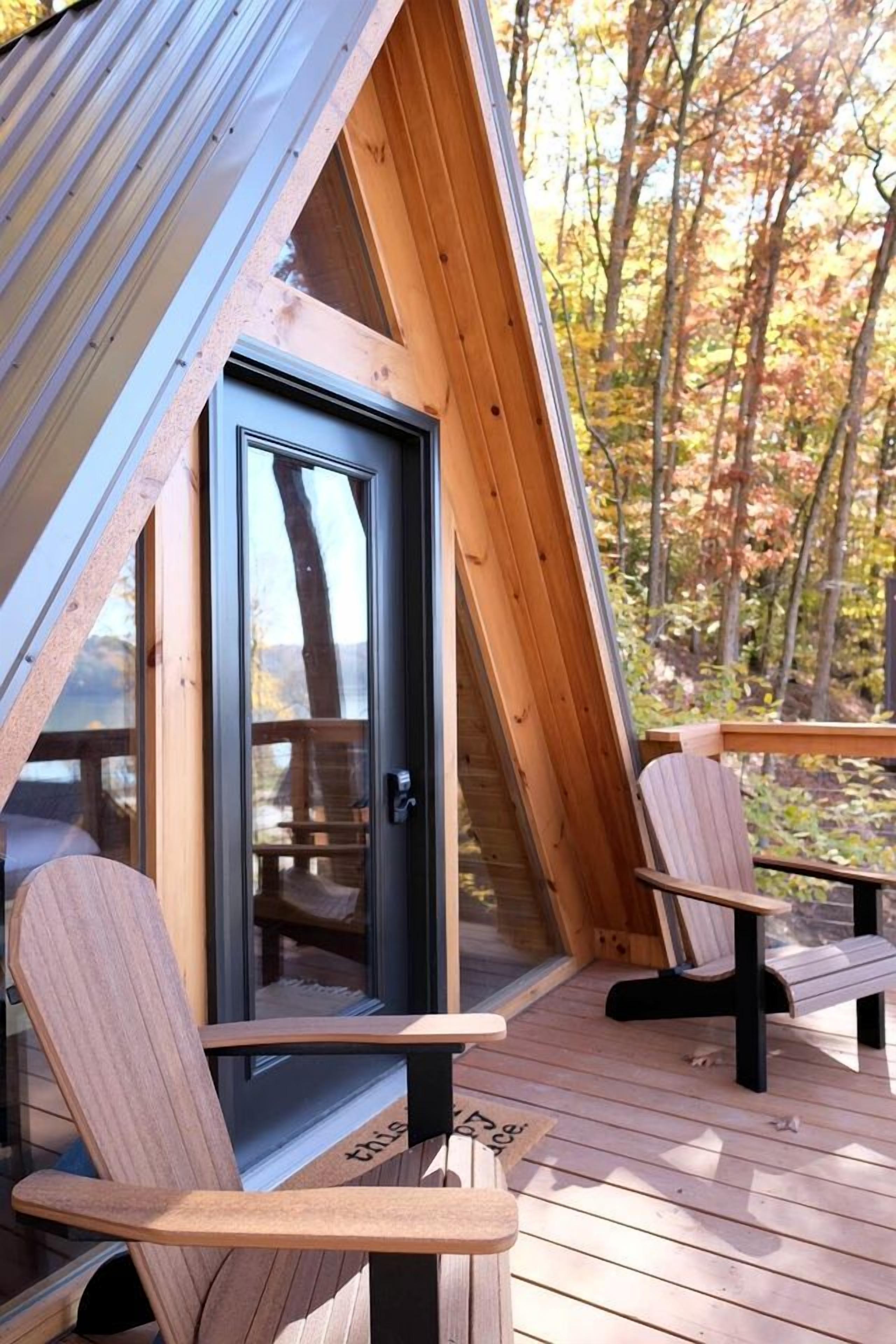 Eine Holzterrasse mit zwei Adirondack-Stühlen, einer Glastür und Blick auf Bäume.