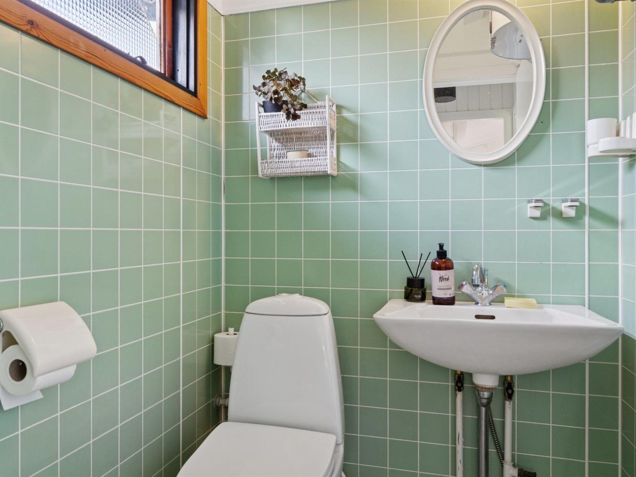 A bathroom with green tiles, a white toilet, a sink with a mirror, and a small plant on a shelf.