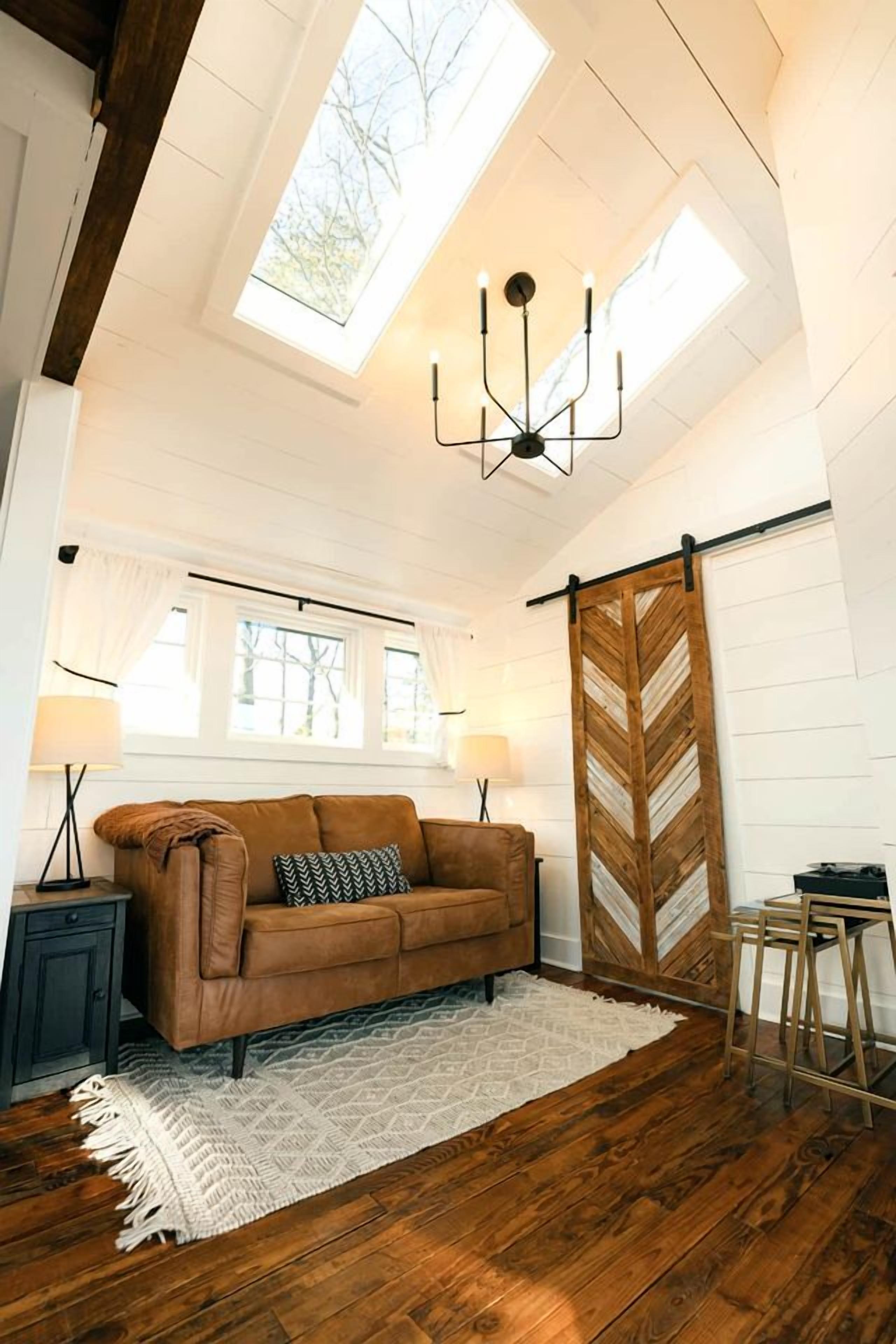 Un ambiente tipo loft con techo alto, vigas de madera, un sofá de cuero marrón, alfombra blanca, panel de pared de madera en patrón chevron y tragaluz.