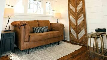 Un ambiente tipo loft con techo alto, vigas de madera, un sofá de cuero marrón, alfombra blanca, panel de pared de madera en patrón chevron y tragaluz.
