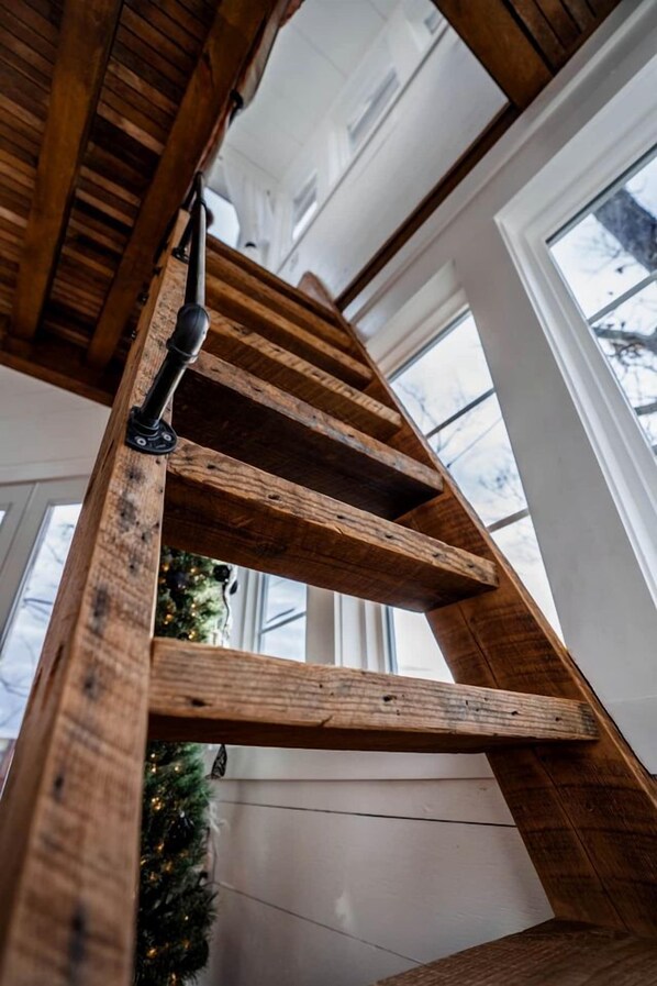 A wooden spiral staircase with a black handrail, leading to a room with large windows and a Christmas tree.