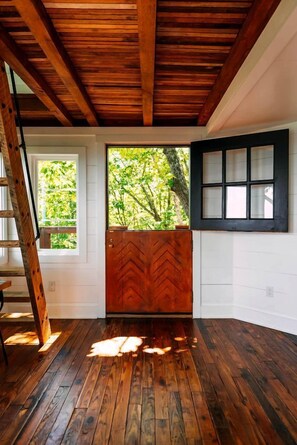 A room with a wooden floor, a herringbone-patterned door, a window with a view of greenery, and a white wall with a black-framed window.
