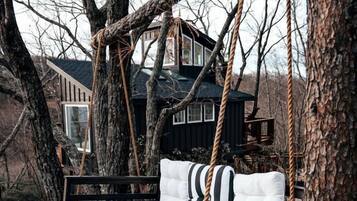 Un lit-balançoire en bois avec un coussin à rayures, entouré d’arbres et avec une maison en arrière-plan.