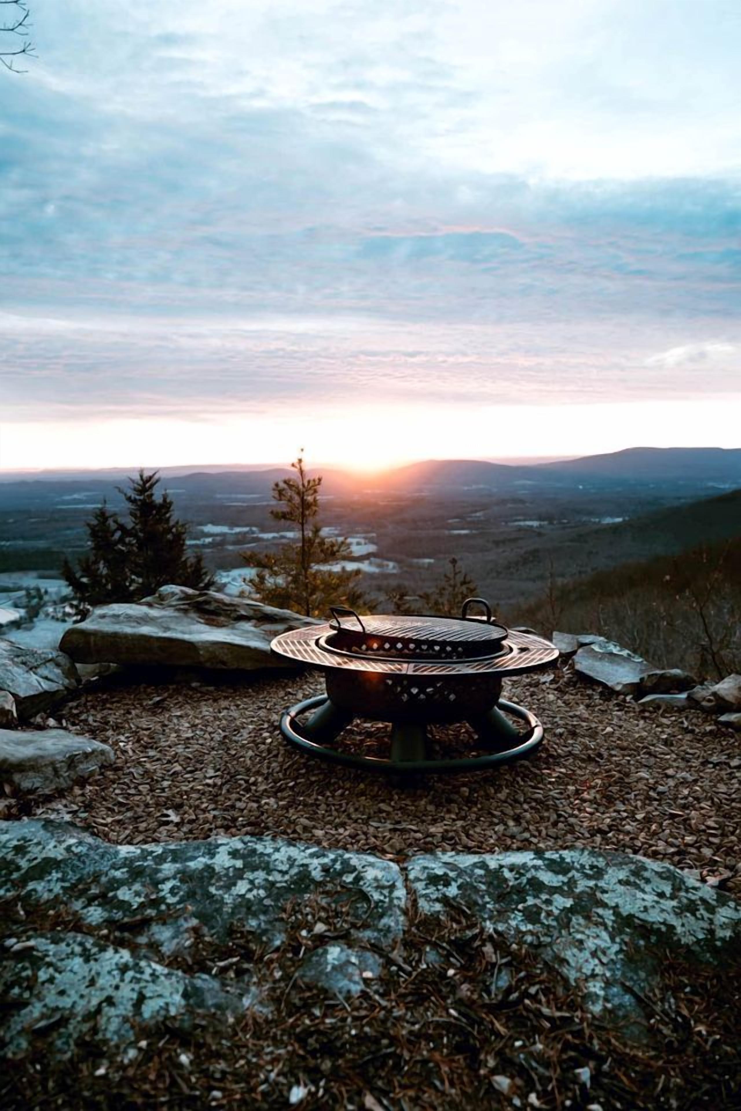A portable grill on a rocky surface with a view of a sunset over a mountainous landscape.