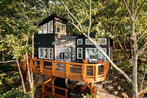 A treehouse with a deck, multiple windows, and a stone base. - Breathtaking Tree House with Jacuzzi and Lofted Interior in Georgia (Rising Fawn)