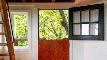 A room with a wooden floor, a herringbone-patterned door, a window with a view of greenery, and a white wall with a black-framed window.