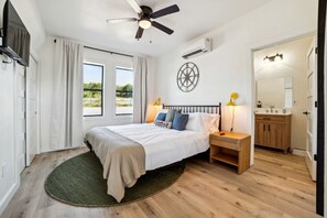 A bedroom with a bed, bedside lamps, a ceiling fan, an air conditioning unit, a round wall clock, and a view of the outdoors through the window. - Elegant Modern A-Frame Surrounded by Nature in Texas (New Braunfels)
