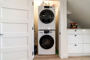 Une buanderie équipée de deux machines à laver à chargement frontal superposées, d’un meuble blanc avec des tiroirs et d’une porte donnant sur une autre pièce.