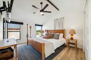 A bedroom with a bed, a desk, a lamp, a ceiling fan, and a window with a view. - Elegant Modern A-Frame Surrounded by Nature in Texas (New Braunfels)