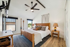 A bedroom with a bed, a desk, a lamp, a ceiling fan, and a window with a view.