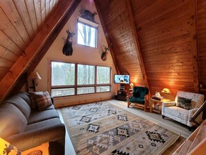 Una habitación estilo loft acogedora, con techo de madera, un ventanal grande, televisor, sofá, alfombra estampada y una silla.