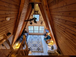 Una habitación con techos altos y un techo inclinado, una ventana grande, un ventilador de techo y una alfombra estampada.