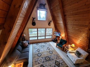 Un acogedor dormitorio en ático con techo inclinado, un ventanal grande, un sofá, una silla, un tapete estampado y decoraciones de animales colgadas en la pared.