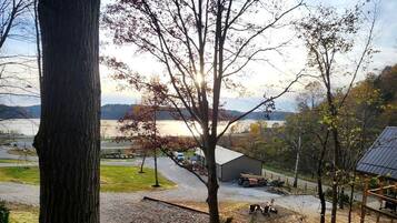 A view of a lakeside area with a wooden deck, a stone fire pit, and a building in the background.