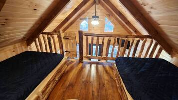 A wooden attic bedroom with a sloped ceiling, a central wooden beam, and a window with a view of trees.