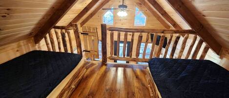 A wooden attic bedroom with a sloped ceiling, a central wooden beam, and a window with a view of trees.