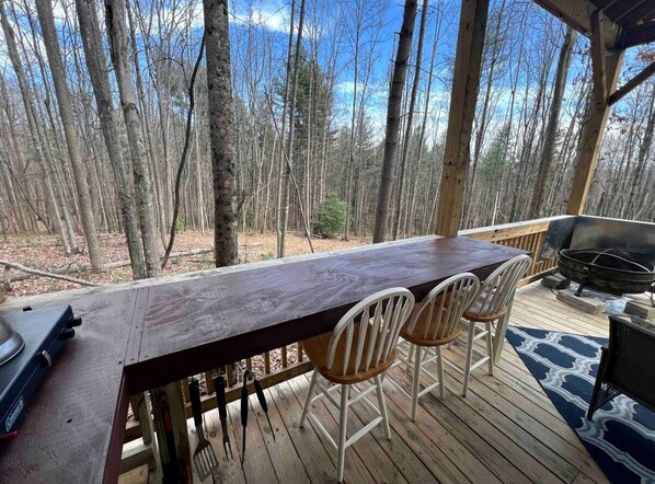 Una terraza de madera con una mesa larga, cuatro sillas, una parrilla y un televisor.