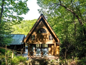 A wooden cabin with a triangular roof, a porch with two chairs, and a metal shed in a forested area.