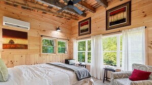 A wooden cabin bedroom with a bed, a chair, a desk, and a window with a view of greenery. - Romantic Riverfront Cabin with Pool Access in Blue Ridge, Georgia (Blue Ridge)