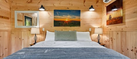 A wooden-paneled bedroom with a bed, two bedside lamps, a wall-mounted air conditioner, and a painting of a sunset.