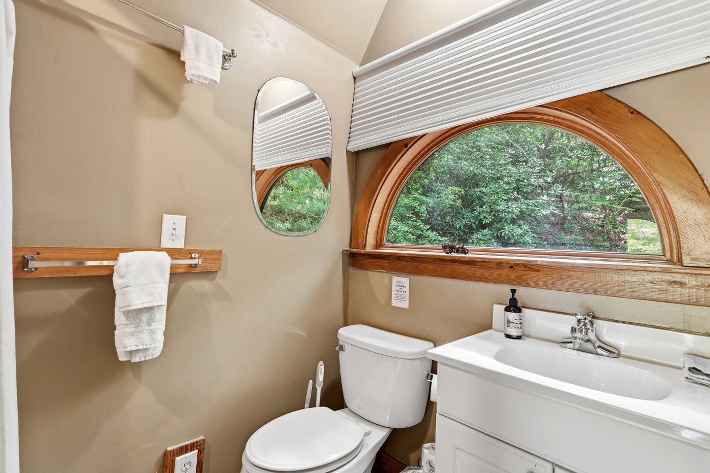 A bathroom with a toilet, sink, mirror, towel rack, and a window with a view of greenery.