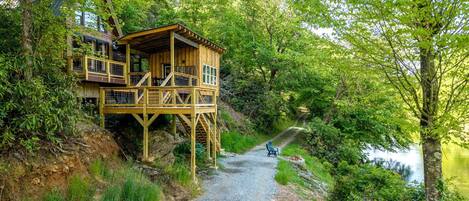 A wooden cabin with a deck, surrounded by trees and a gravel path leading to a body of water.