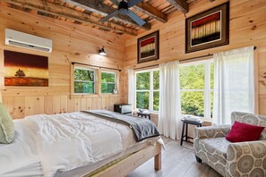 A wooden cabin bedroom with a bed, a chair, a desk, and a window with a view of greenery.