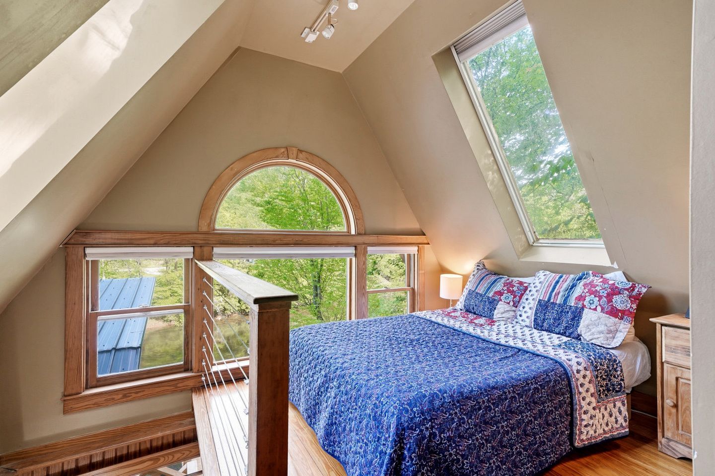 Ein Schlafzimmer mit schrägen Wänden, einem Bett mit blauer Steppdecke, einem Nachttisch und einem Fenster mit Blick auf Grünflächen.