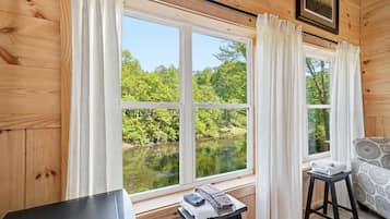 A cabin with wooden walls, a window with a view of a lake surrounded by trees, a small table with towels, and a patterned sofa.