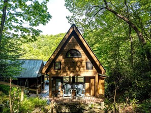 A wooden cabin with a triangular roof, a porch with two chairs, and a metal shed in a forested area.