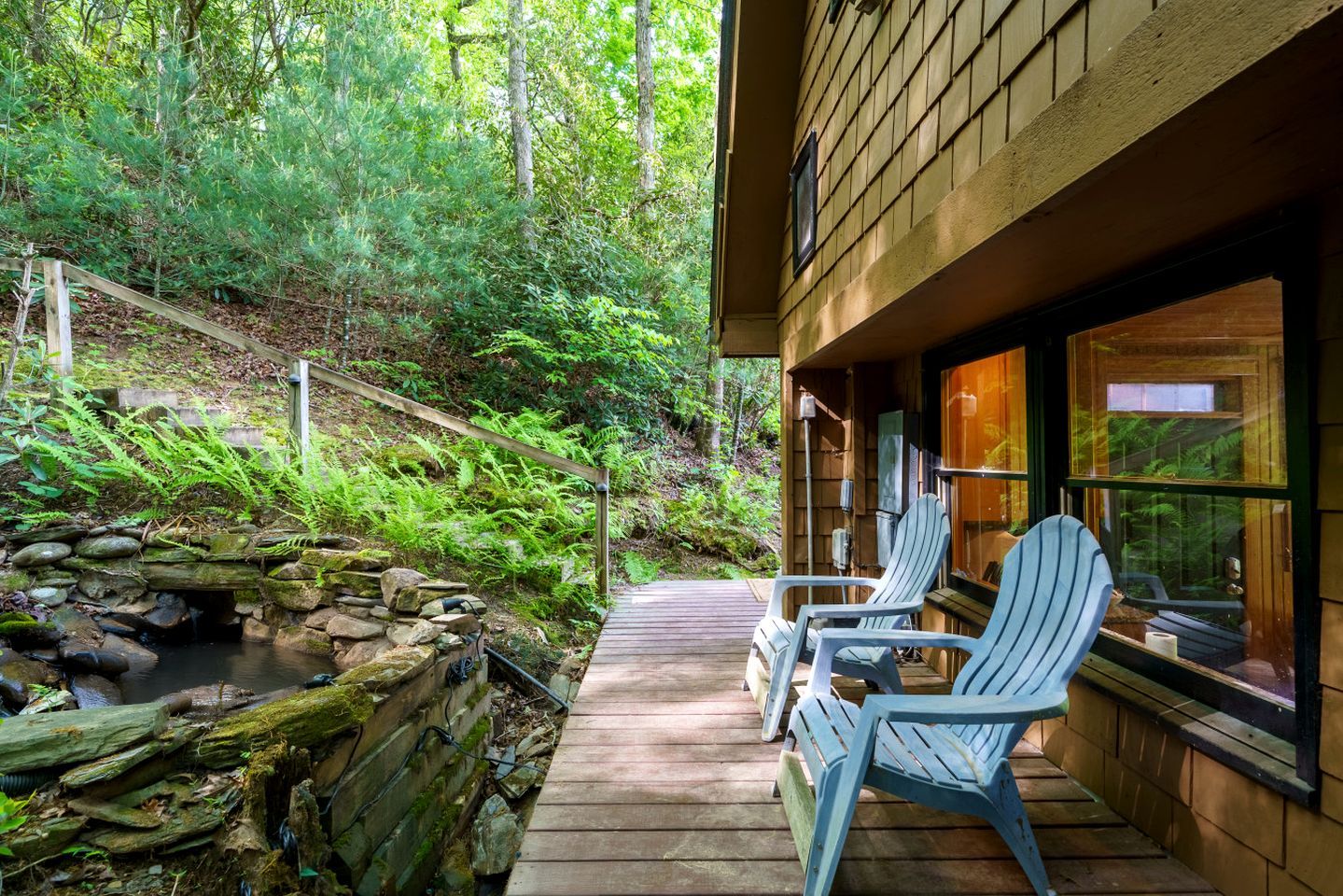 Une terrasse en bois avec des fauteuils Adirondack, un mur de pierres et un petit ruisseau.