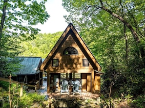A wooden cabin with a triangular roof, a porch with two chairs, and a metal shed in a forested area.