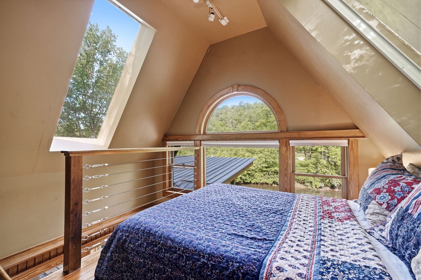 Ein Schlafzimmer mit Oberlicht, einem Bett mit gemusterten Betttextilien, einem hölzernen Kopfteil und Blick auf Bäume durch große Fenster.