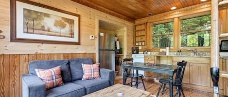 A cozy cabin interior with a sofa, coffee table, dining table, chairs, kitchen area, and a framed picture on the wall.
