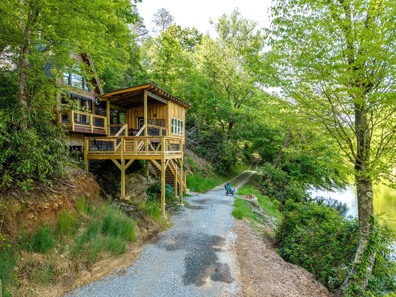 A wooden cabin with a deck, surrounded by trees and a gravel path leading to a body of water.