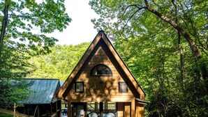 A wooden cabin with a triangular roof, a porch with two chairs, and a metal shed in a forested area.
