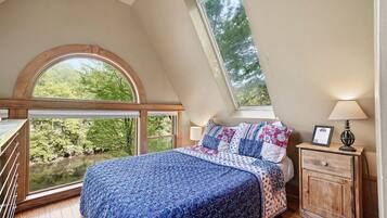 Un dormitorio con un ventanal grande, una cama con ropa de cama estampada, una mesita de noche con lámpara y vistas al exterior con vegetación.