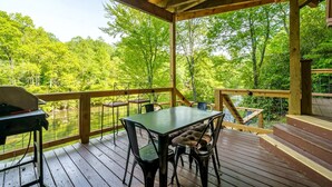 A covered outdoor deck with a table and chairs, overlooking a forested area. - Romantic Riverfront Cabin with Pool Access in Blue Ridge, Georgia (Blue Ridge)