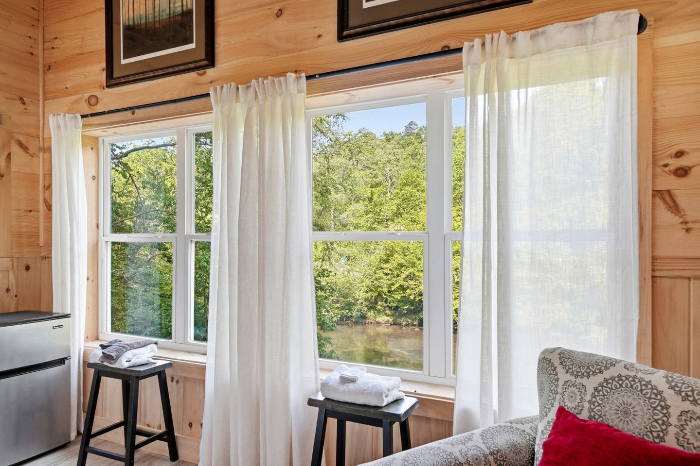 Une cabane aux murs en bois, avec un coin cuisine, un canapé agrémenté d’un coussin à motifs et une vue sur une zone boisée, visible à travers de grandes fenêtres.