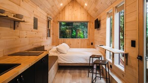 A compact cabin with a bed, a small kitchen, and a window with a forest view.