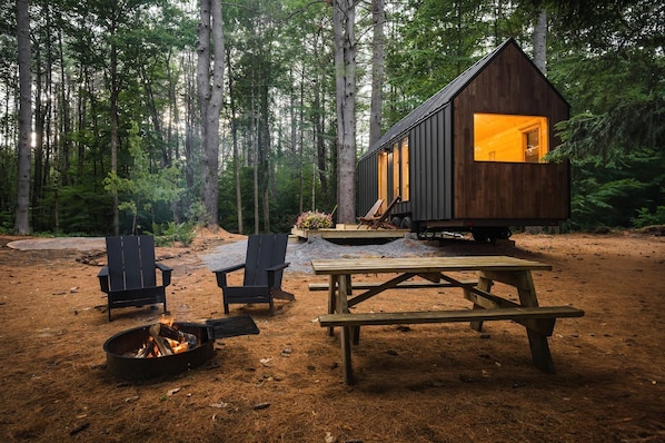 A cabin with a fire pit, two chairs, and a picnic table in a forest setting.