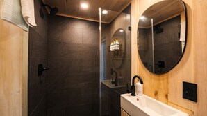 A modern bathroom with dark tiles, a wooden ceiling, a glass shower enclosure, a round mirror, and a white sink.