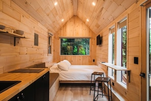 A compact cabin with a bed, a small kitchen, and a window with a forest view.