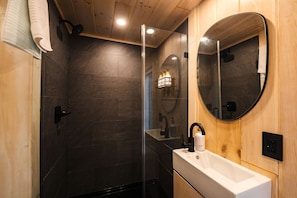 A modern bathroom with dark tiles, a wooden ceiling, a glass shower enclosure, a round mirror, and a white sink.