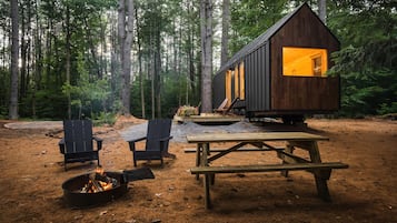 A cabin with a fire pit, two chairs, and a picnic table in a forest setting.