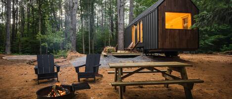 Une cabane avec un foyer, deux chaises et une table de pique-nique, le tout situé dans un cadre forestier.