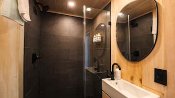 A modern bathroom with dark tiles, a wooden ceiling, a glass shower enclosure, a round mirror, and a white sink.